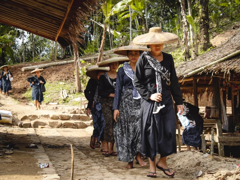 Foto grup Suku Baduy dalam pakaian tradisional hitam berjalan melalui hutan lebat di Banten, menunjukkan kehidupan sehari-hari mereka yang erat dengan alam